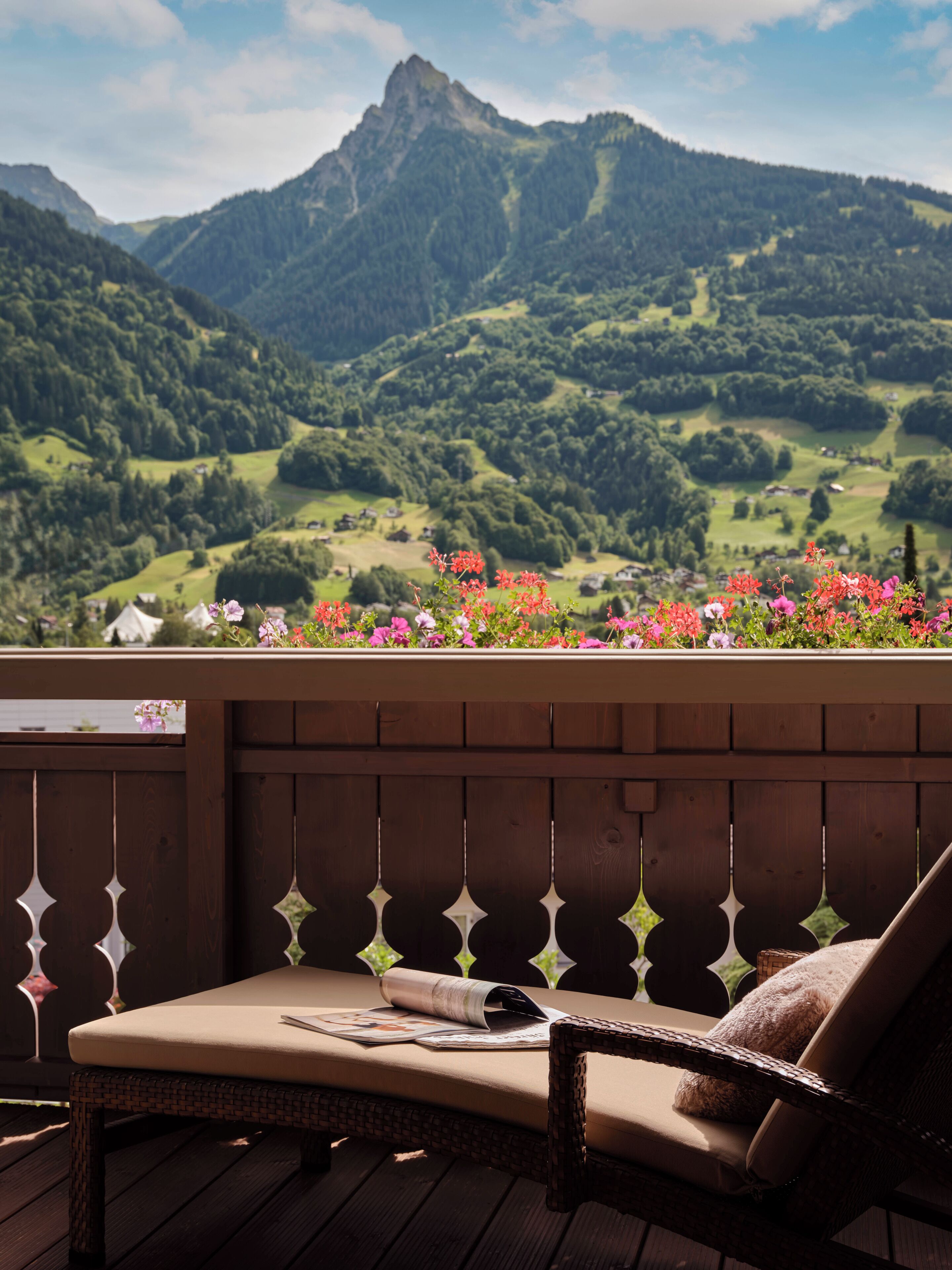 Una terraza de madera con una silla de mimbre y una mesa, con vistas a un paisaje montañoso con vegetación y un cantero de flores.