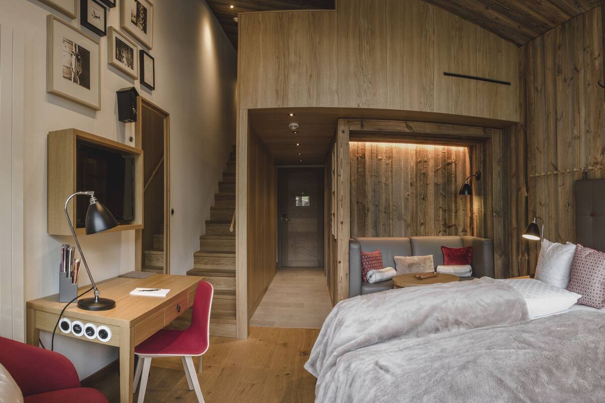 A modern bedroom with a wooden ceiling, a desk with a chair, a bed with a headboard, and a staircase leading to another room.