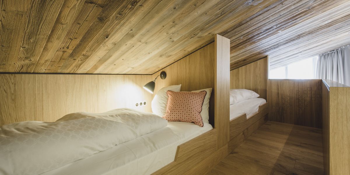 A wooden cabin bedroom with a bunk bed, a small desk, and a window with curtains.