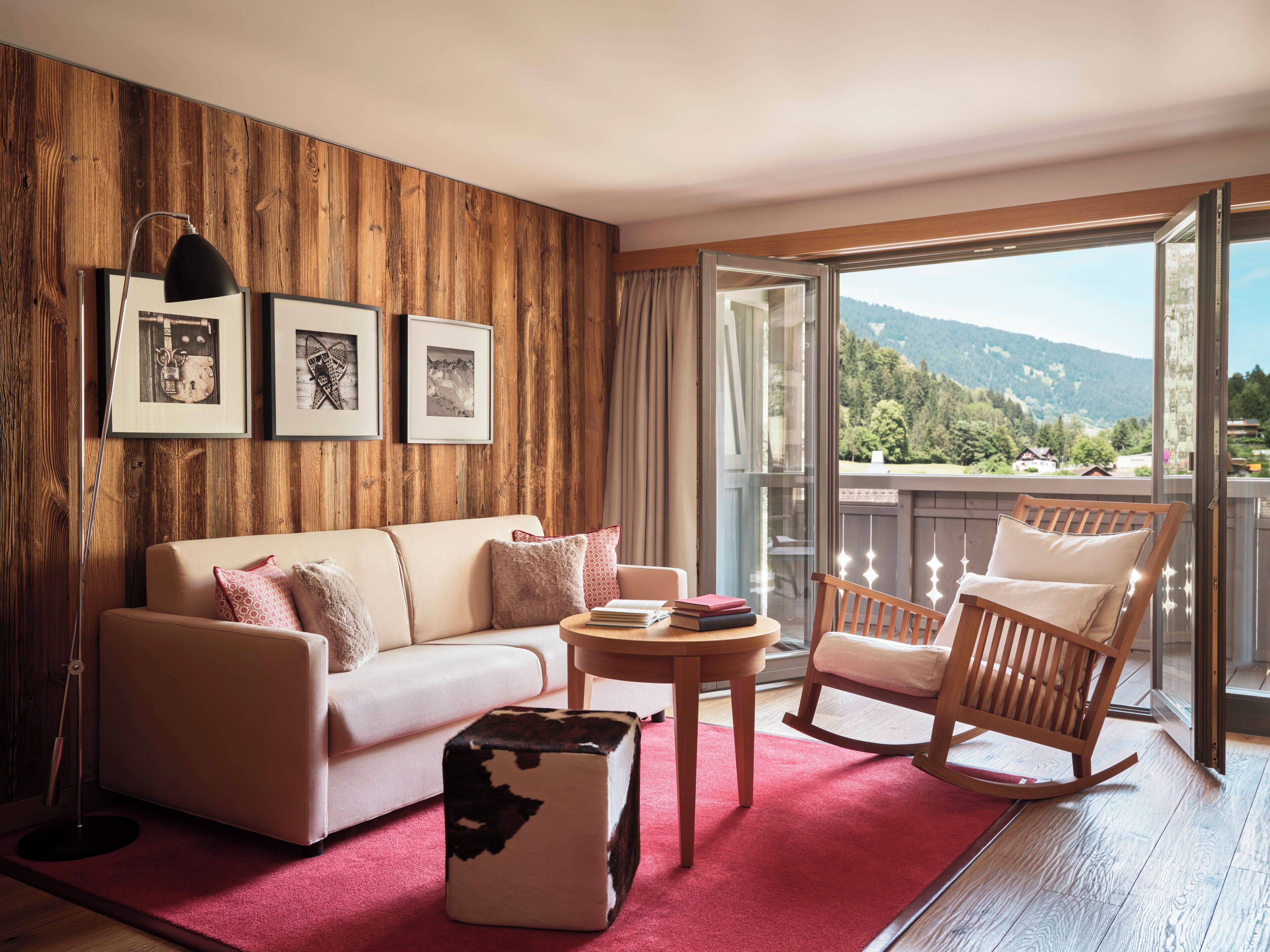 A room with a wooden wall, a sofa, a rocking chair, a small table, and a red rug. There are framed pictures on the wall and a view of the outdoors.