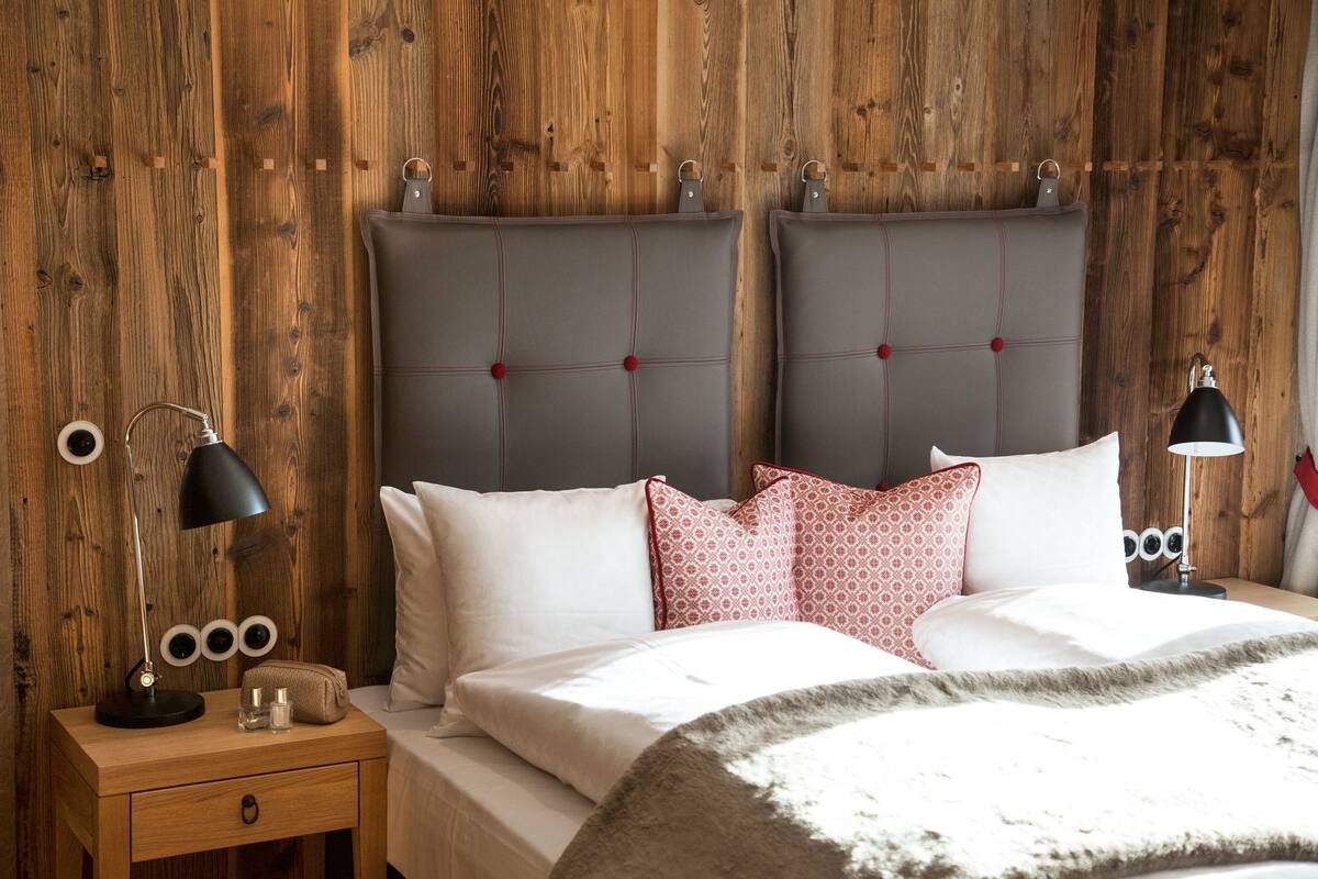 A bedroom with two beds, wooden panel walls, a nightstand with a lamp, and a bedside table with a lamp and a small bag.