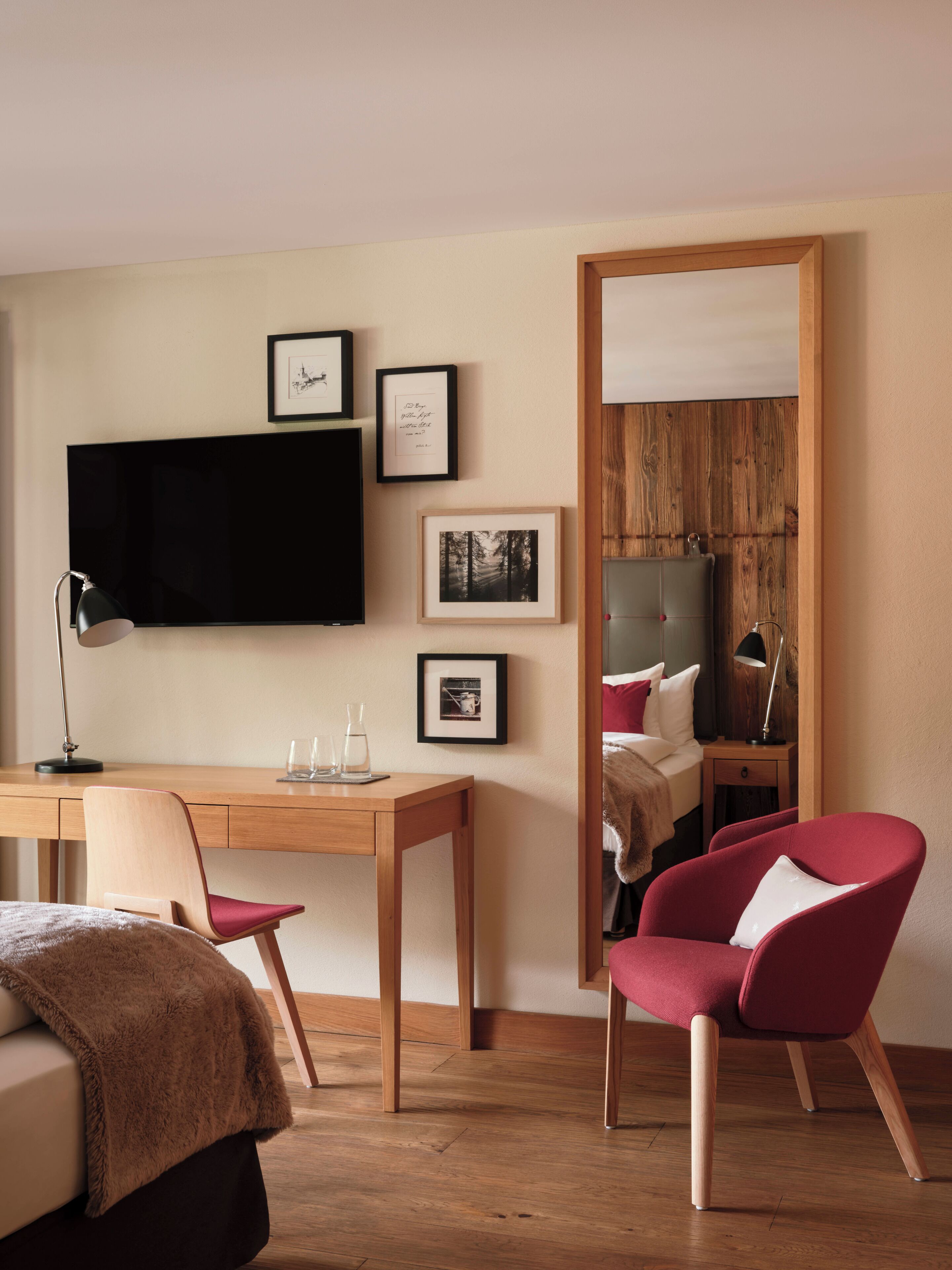 Habitación de hotel con escritorio de madera, una silla, cama con sillón rojo, televisor de pantalla plana y cuadros en la pared.