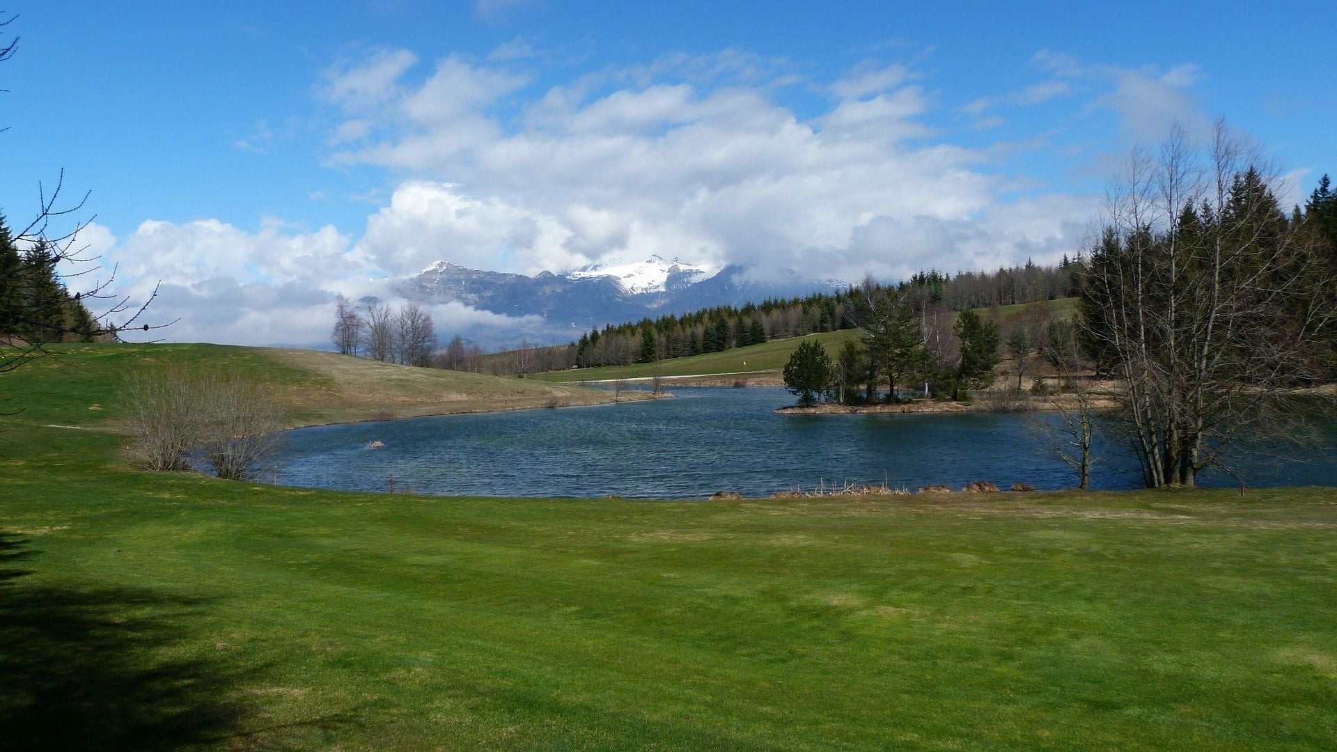 Un paysage paisible au bord d’un lac, avec un premier plan verdoyant, un lac calme et, en arrière-plan, des montagnes enneigées sous un ciel partiellement nuageux.