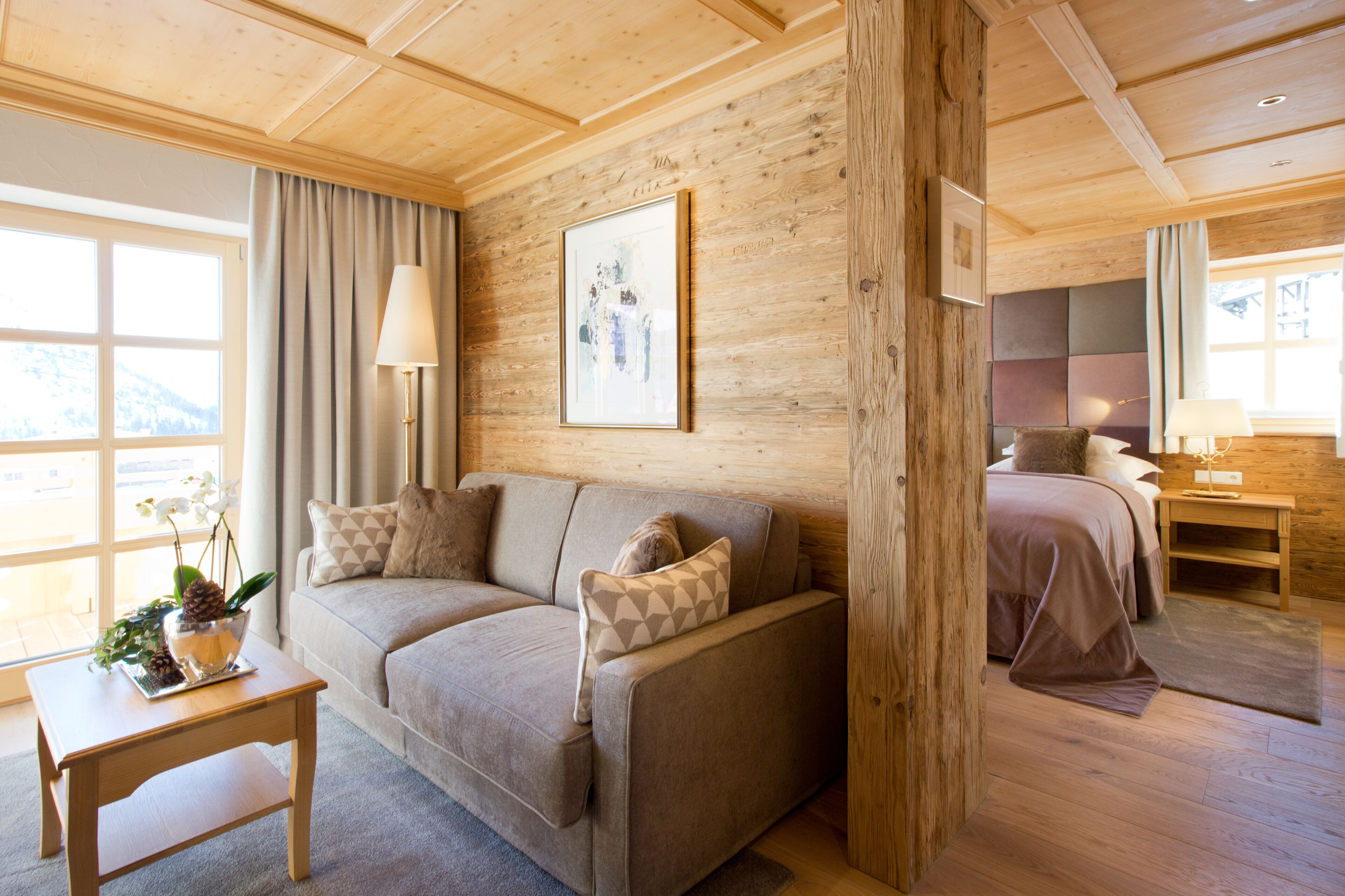 A cozy room with a wooden ceiling, a sofa with patterned cushions, a bedside table with a lamp, and a bed with a headboard.