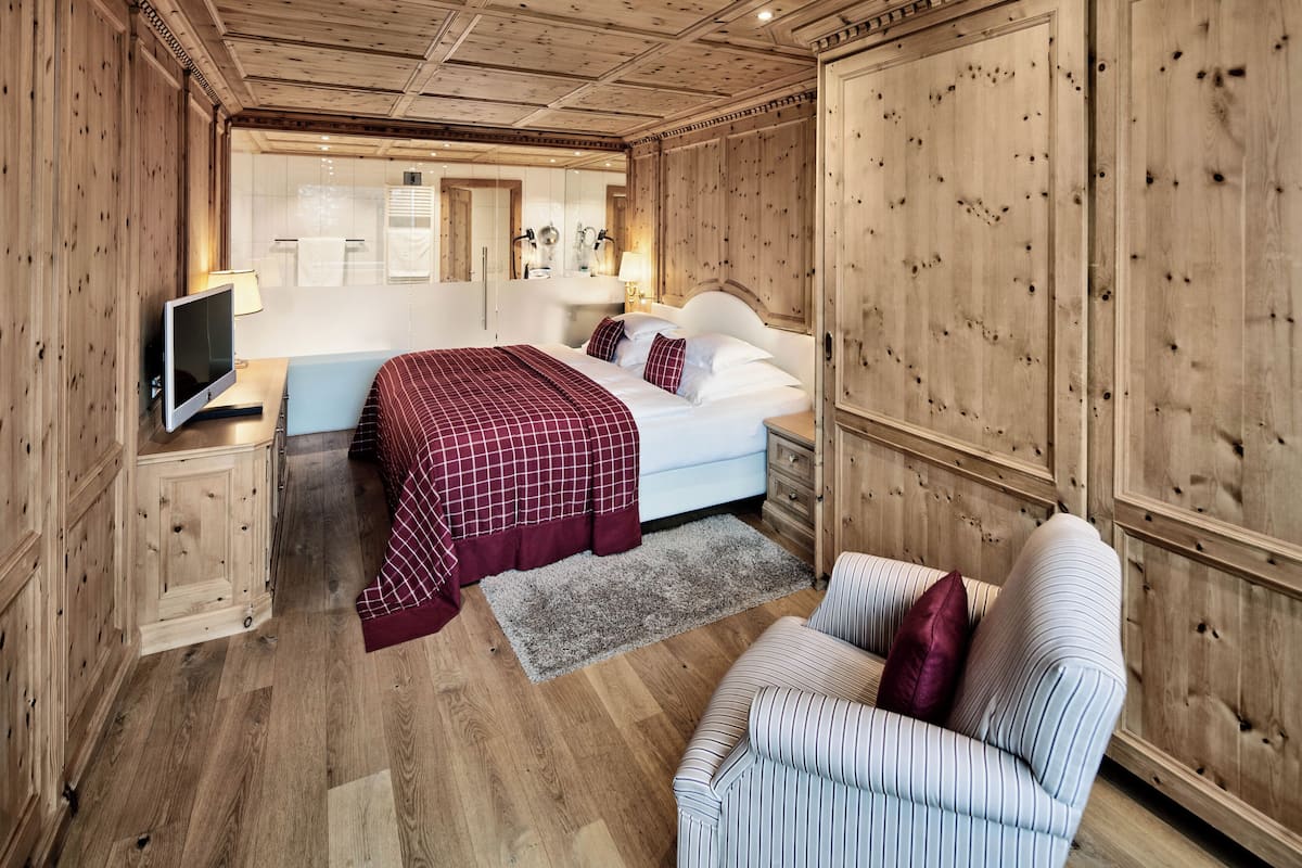 A bedroom with a bed, a TV, a bedside table, a chair, and wooden paneling on the walls.