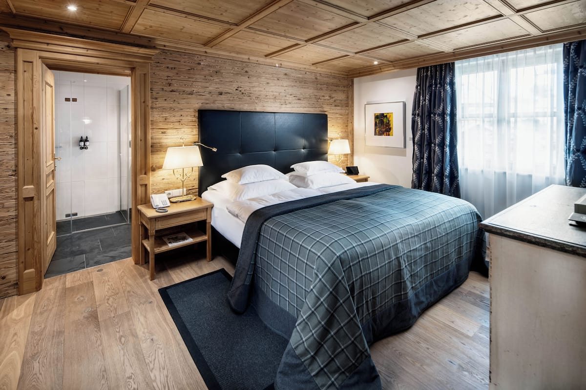 A hotel room with a wooden ceiling, a large bed with a dark headboard, two bedside tables with lamps, a small desk, and a window with curtains.