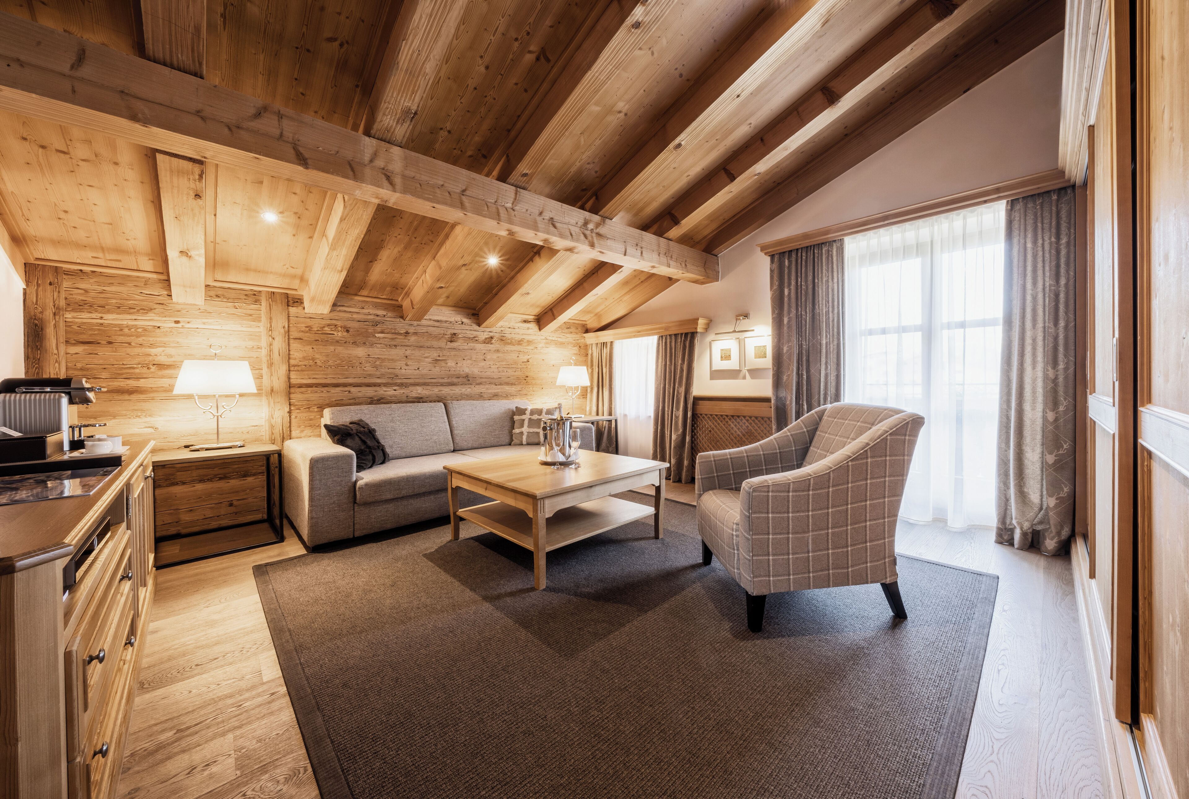 Een gezellige woonkamer met een houten plafond, een bank, een geruite fauteuil, een salontafel en een lamp.