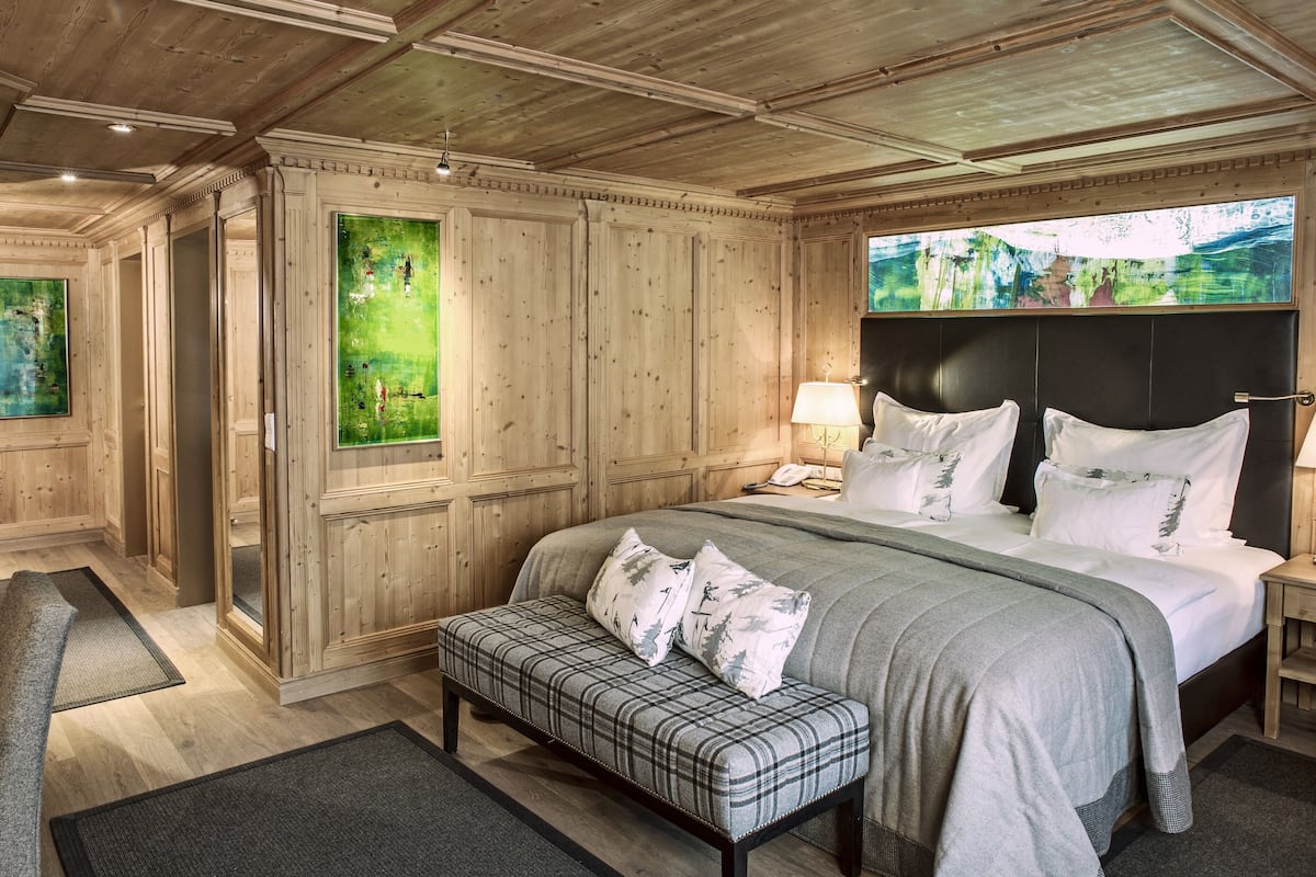 A bedroom with a large bed, wooden paneling, a bench, and a view of greenery through a window.