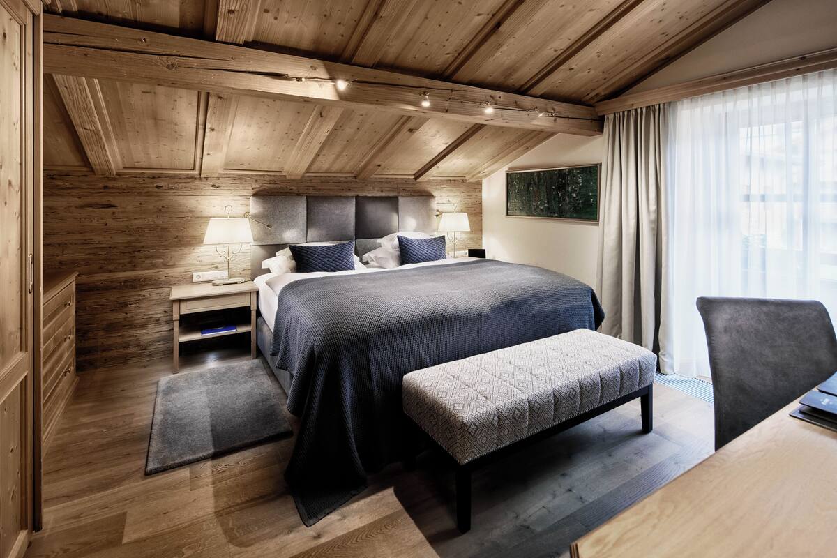 A modern bedroom with a wooden ceiling, a large bed with a dark blue bedspread, a bedside table with lamps, and a view of the outdoors through a window.