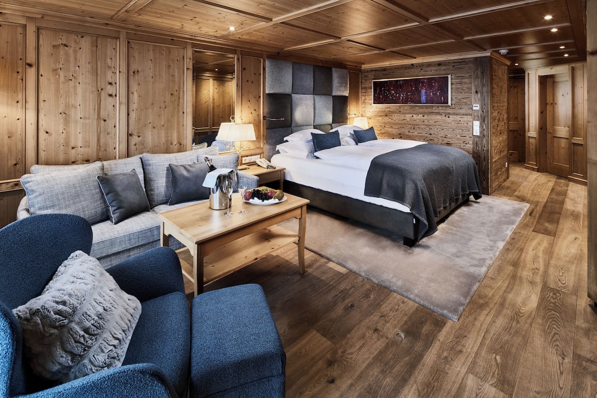 A modern hotel room with a wooden ceiling, a large bed with a grey headboard, a sofa, a wooden table, and a nightstand with a lamp.