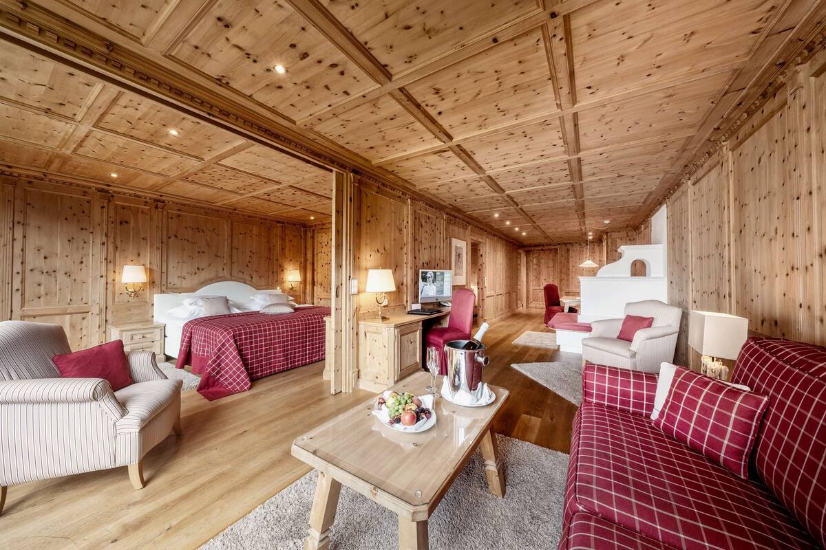 A wooden cabin interior with a bed, sofa, armchair, and a table with fruit and a champagne bucket.