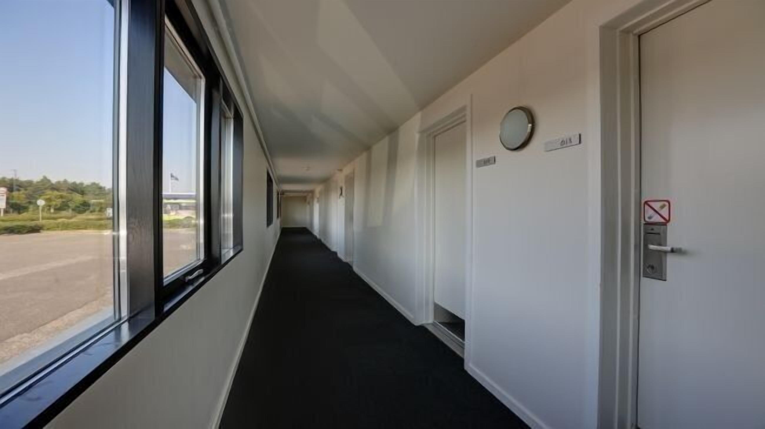 Un long couloir étroit, doté d’un tapis noir, de murs blancs et d’une fenêtre sur le côté gauche. Des portes se trouvent sur le côté droit, dont une porte sur laquelle est apposé un panneau « interdiction de fumer ».