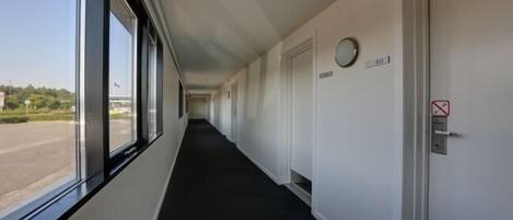 Un long couloir étroit, doté d’un tapis noir, de murs blancs et d’une fenêtre sur le côté gauche. Des portes se trouvent sur le côté droit, dont une porte sur laquelle est apposé un panneau « interdiction de fumer ».