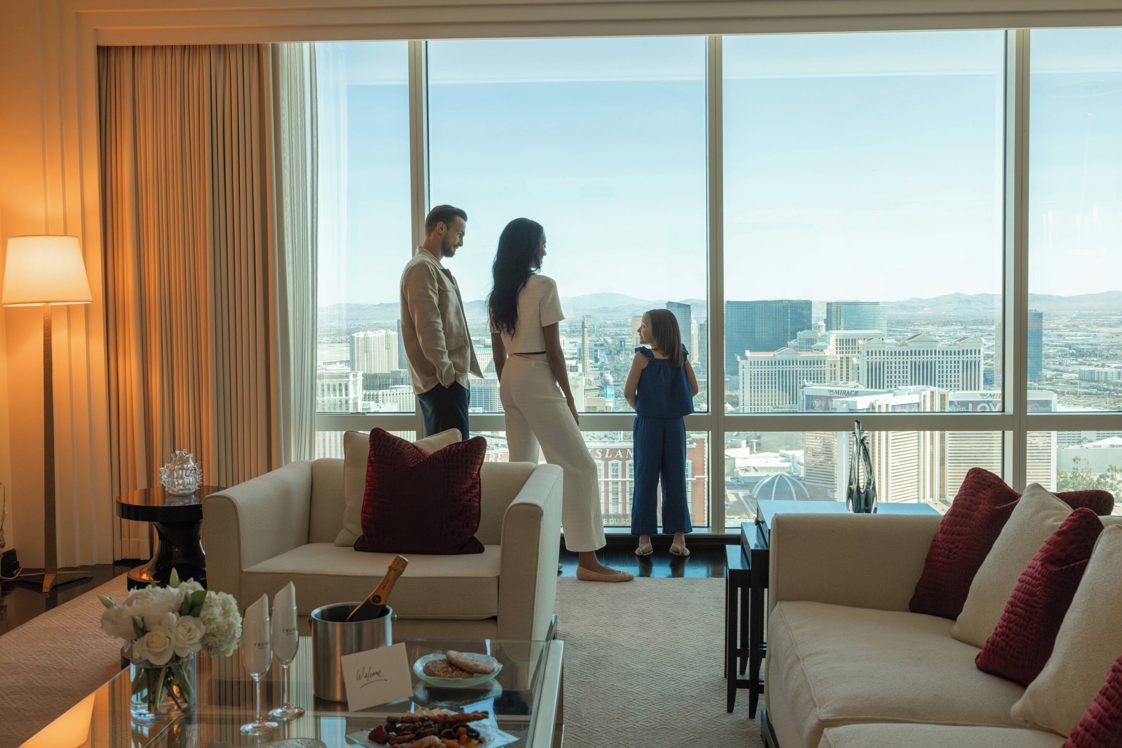 A hotel room with a large window offering a city view, a sofa, a coffee table with a cake and glasses, and a vase with flowers.