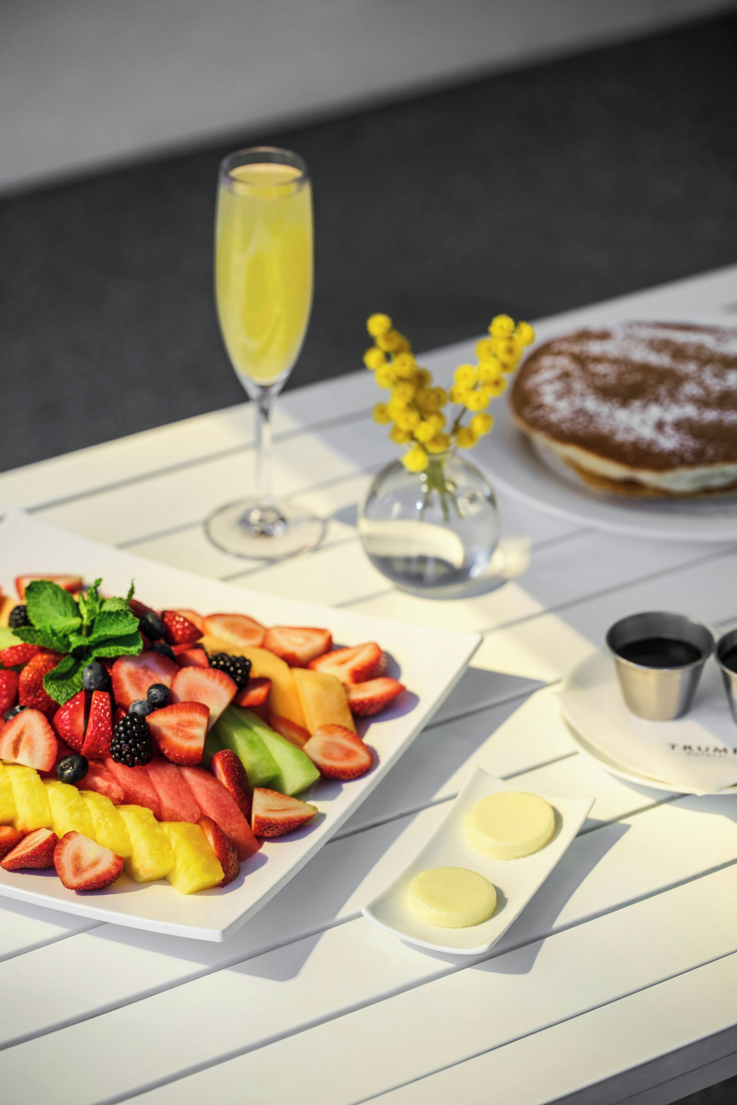 A table with a fruit platter, a glass of yellow beverage, a pastry, and two small cups.
