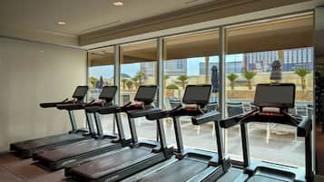 A row of treadmills with digital screens, large windows with a view of palm trees and buildings, and a ceiling with recessed lighting.