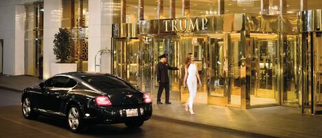 A building entrance with a glass facade, a sign reading 'TRUMP', and a black car parked outside.