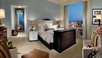 A hotel room with a large bed, a nightstand, a dresser, a sitting area with a chair and ottoman, and a view of the cityscape.