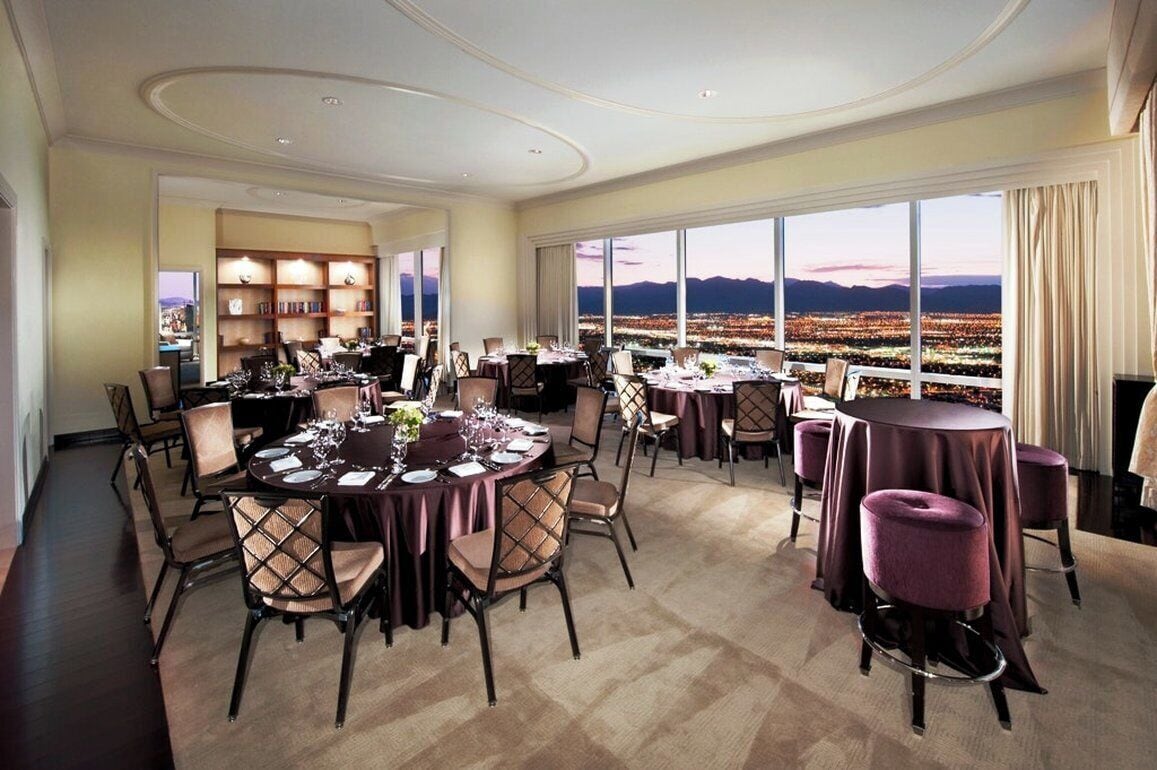 A dining area with round tables and chairs, a bookshelf, and a large window with a cityscape view.