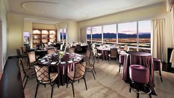 A dining area with round tables and chairs, a bookshelf, and a large window with a cityscape view.