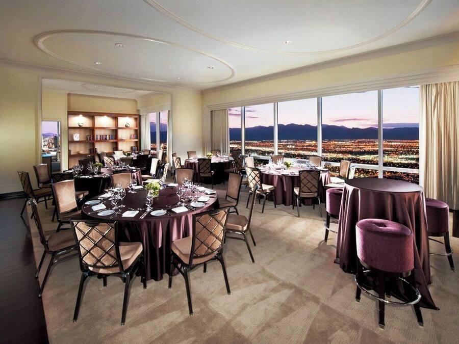 A dining area with round tables and chairs, a bookshelf, and a large window with a cityscape view.