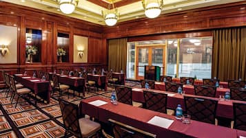 Un salón de conferencias con varias mesas y sillas, paneles de madera y ventanas grandes con cortinas.