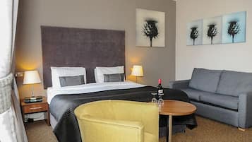 A hotel room with a bed, a sofa, a small table with a bottle, and wall art.