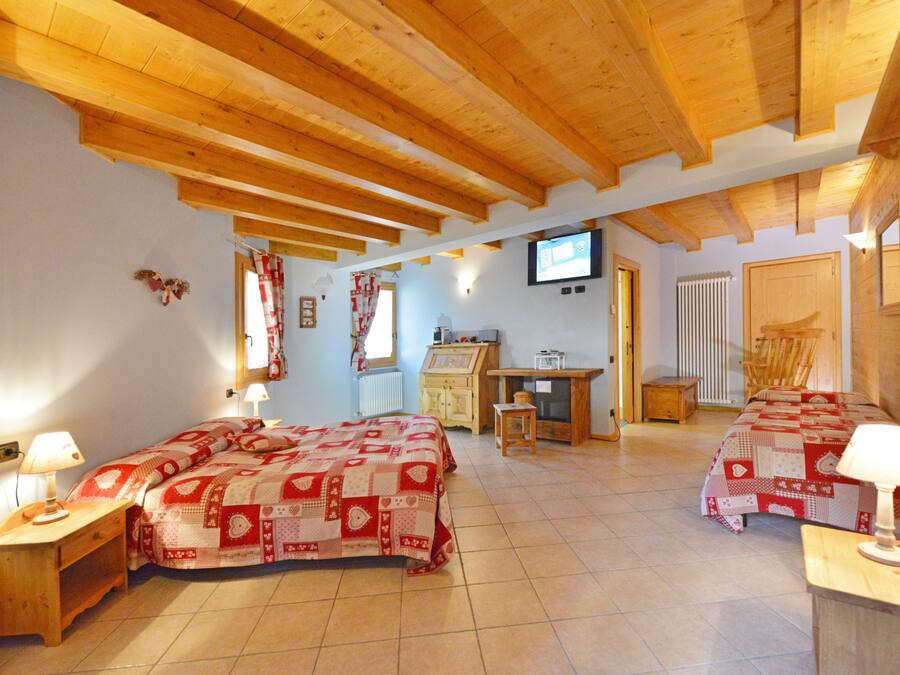 A cozy bedroom with a wooden ceiling, a bed with a red and white quilt, a desk with a computer, and a television on the wall.