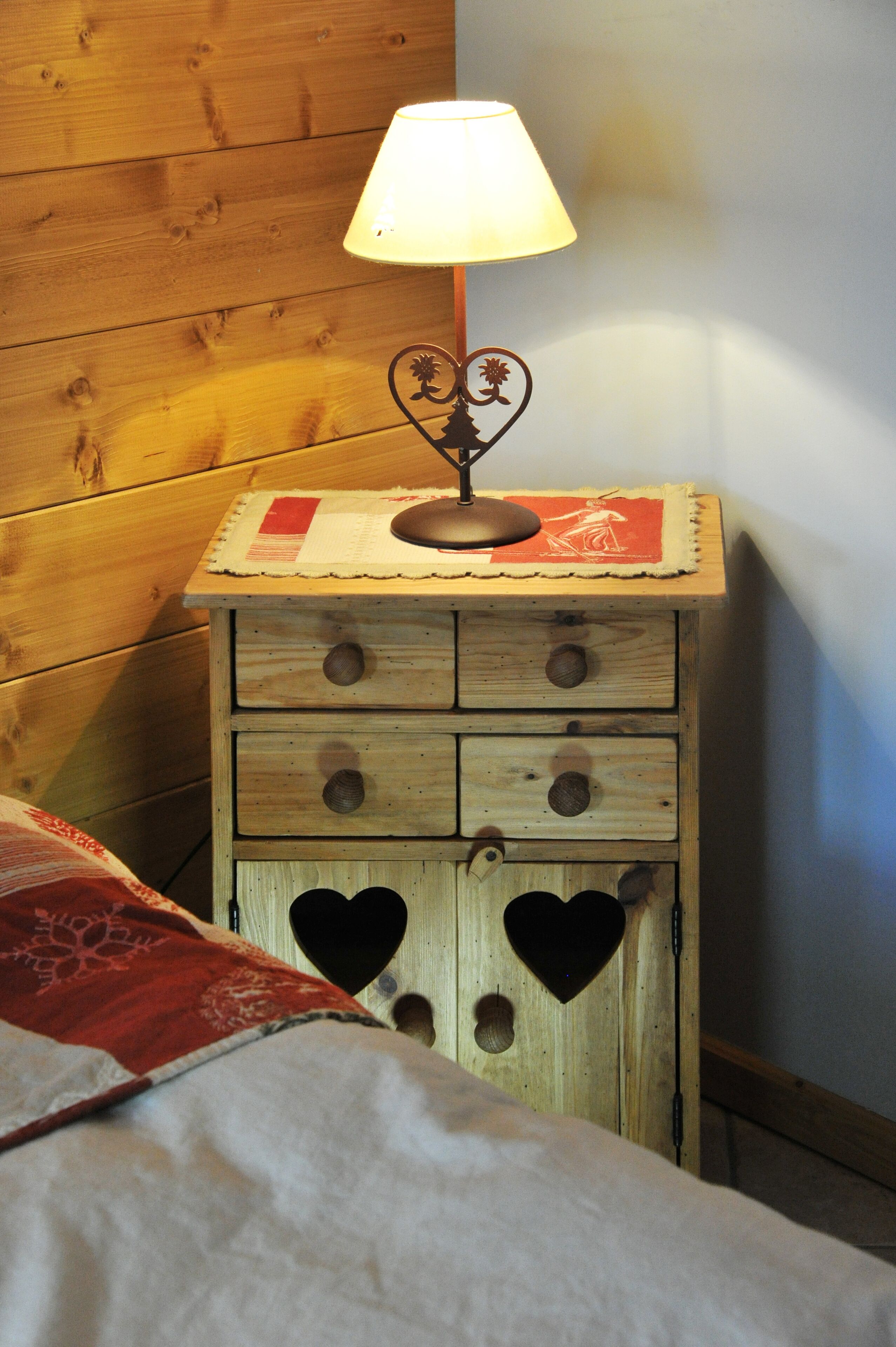 Une table de chevet en bois, avec une lampe, un set de table rouge et blanc, et des découpes en forme de cœur sur le devant.