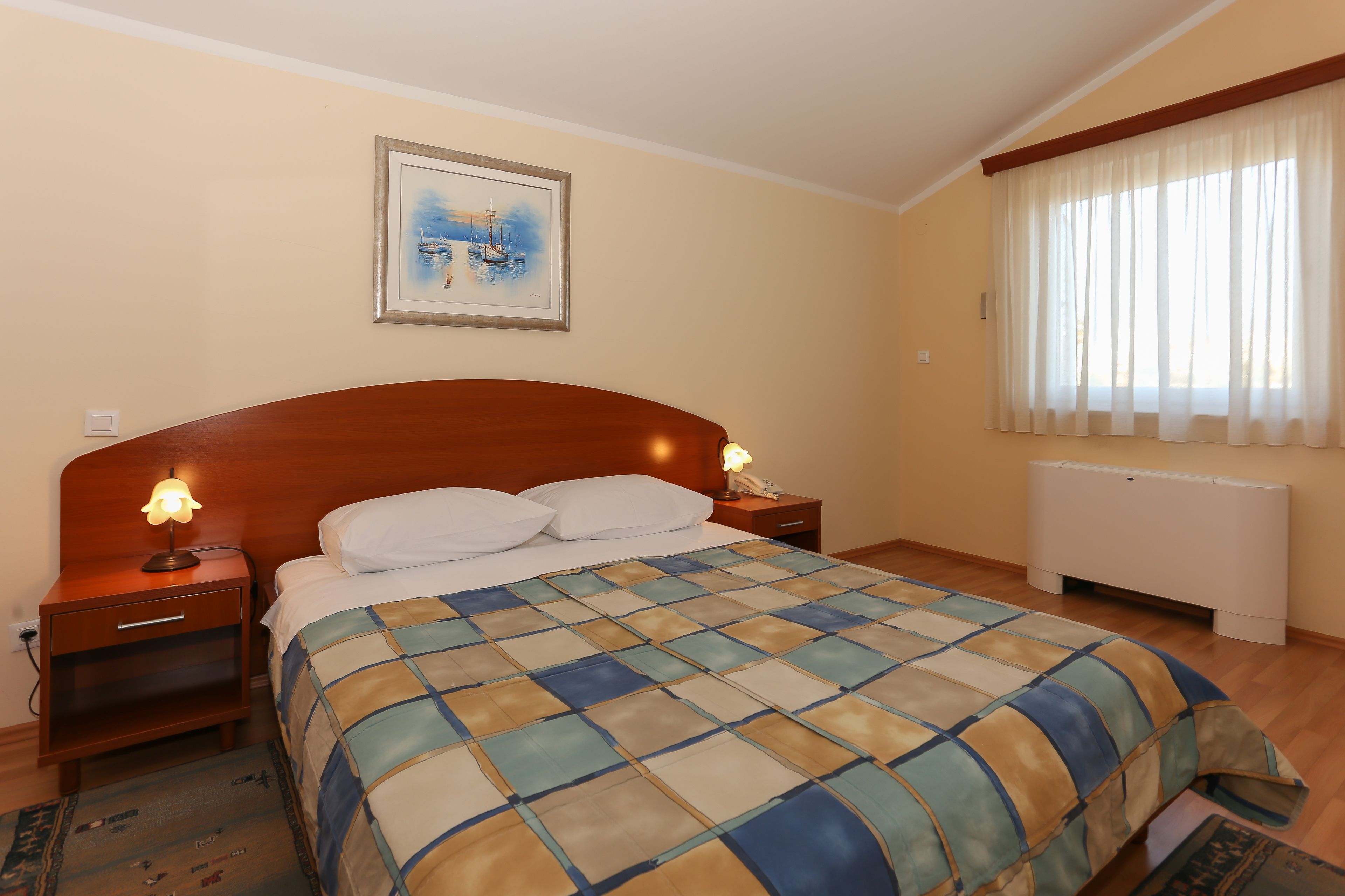 A bedroom with a wooden headboard, a bed with a checkered bedspread, two bedside lamps, a nightstand, a framed picture on the wall, and a window with sheer curtains.