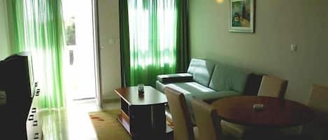 A living room with a sofa, a coffee table, and a TV. There is a door leading outside and a painting on the wall.