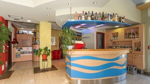 A hotel lobby with a reception desk, a bar area with various liquor bottles, a red chair, and a potted plant. - Aparthotel Astoria (Seget)