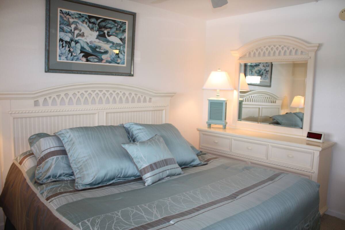Ein Schlafzimmer mit Bett, Nachttisch, Lampe, Kommode und einem gerahmten Bild an der Wand.