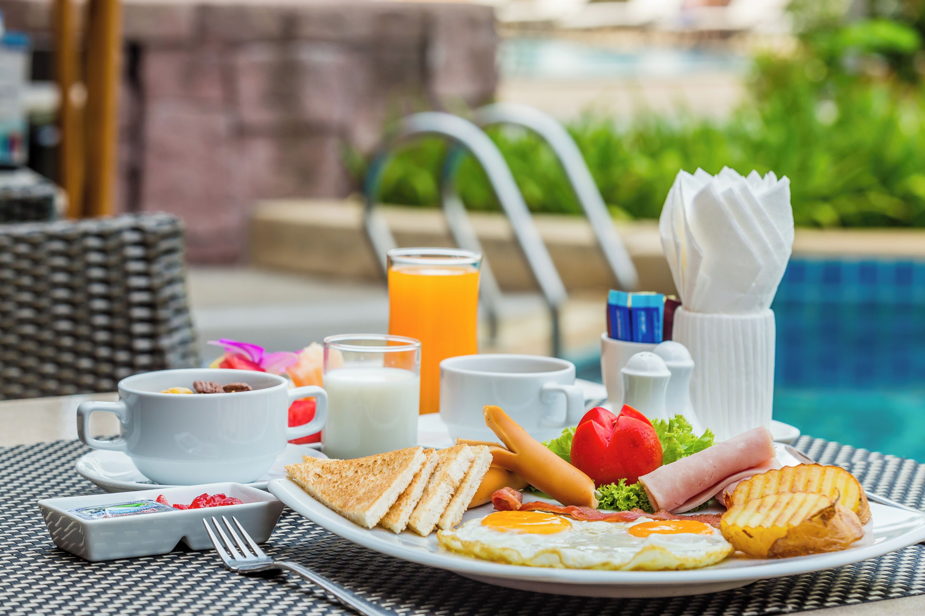 Une table dressée pour le petit-déjeuner avec une assiette d’œufs, des toasts, du bacon et du jambon, un verre de jus d’orange et une tasse de café.