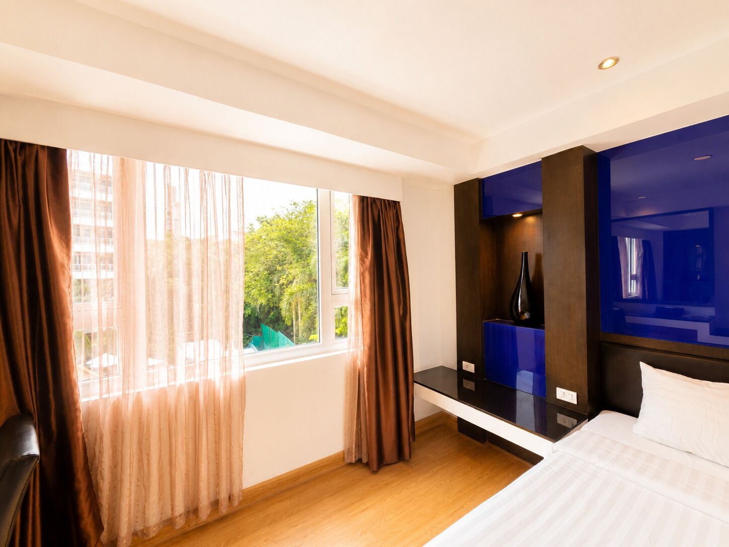A hotel room with a large window, a bed, a bedside table, and a blue accent wall.
