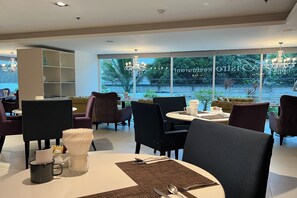 A modern dining area with round tables, chairs, and a view of a garden through large windows. - Nova Suites Pattaya by Compass Hospitality (Pattaya)