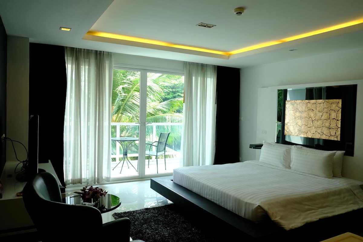Una habitación de hotel moderna con una cama grande, una mesa de vidrio con un jarrón de flores, un televisor y vistas al exterior a través de amplias ventanas.