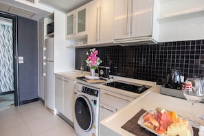 A modern kitchen with white cabinetry, a built-in washing machine, a black tiled backsplash, and a fruit platter on the counter. - Nova Suites Pattaya by Compass Hospitality (Pattaya)