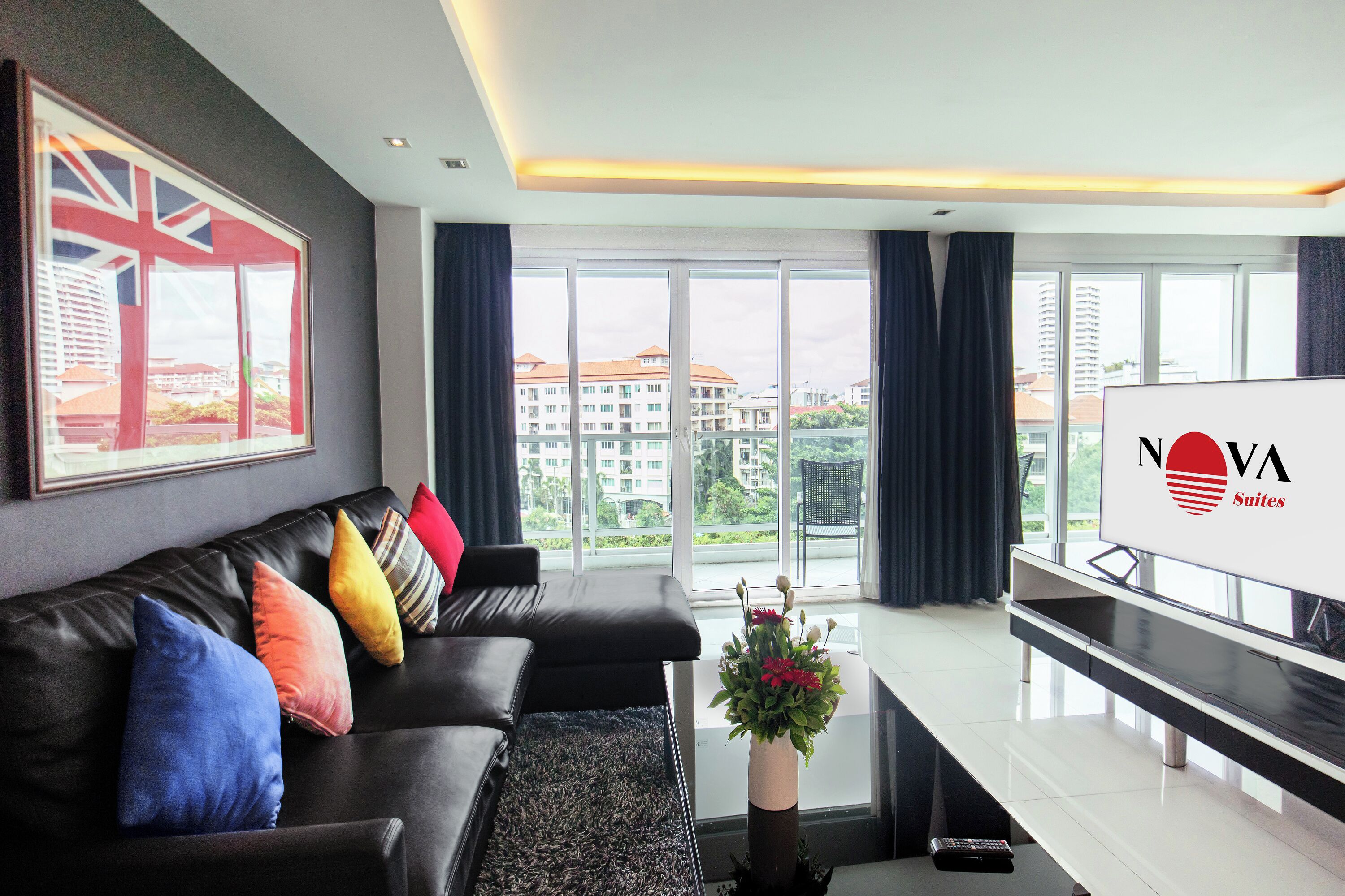 A modern living room with a black leather sofa, colorful cushions, a coffee table with a vase of flowers, and a large window offering a city view.