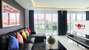 A modern living room with a black leather sofa, colorful cushions, a coffee table with a vase of flowers, and a large window offering a city view.