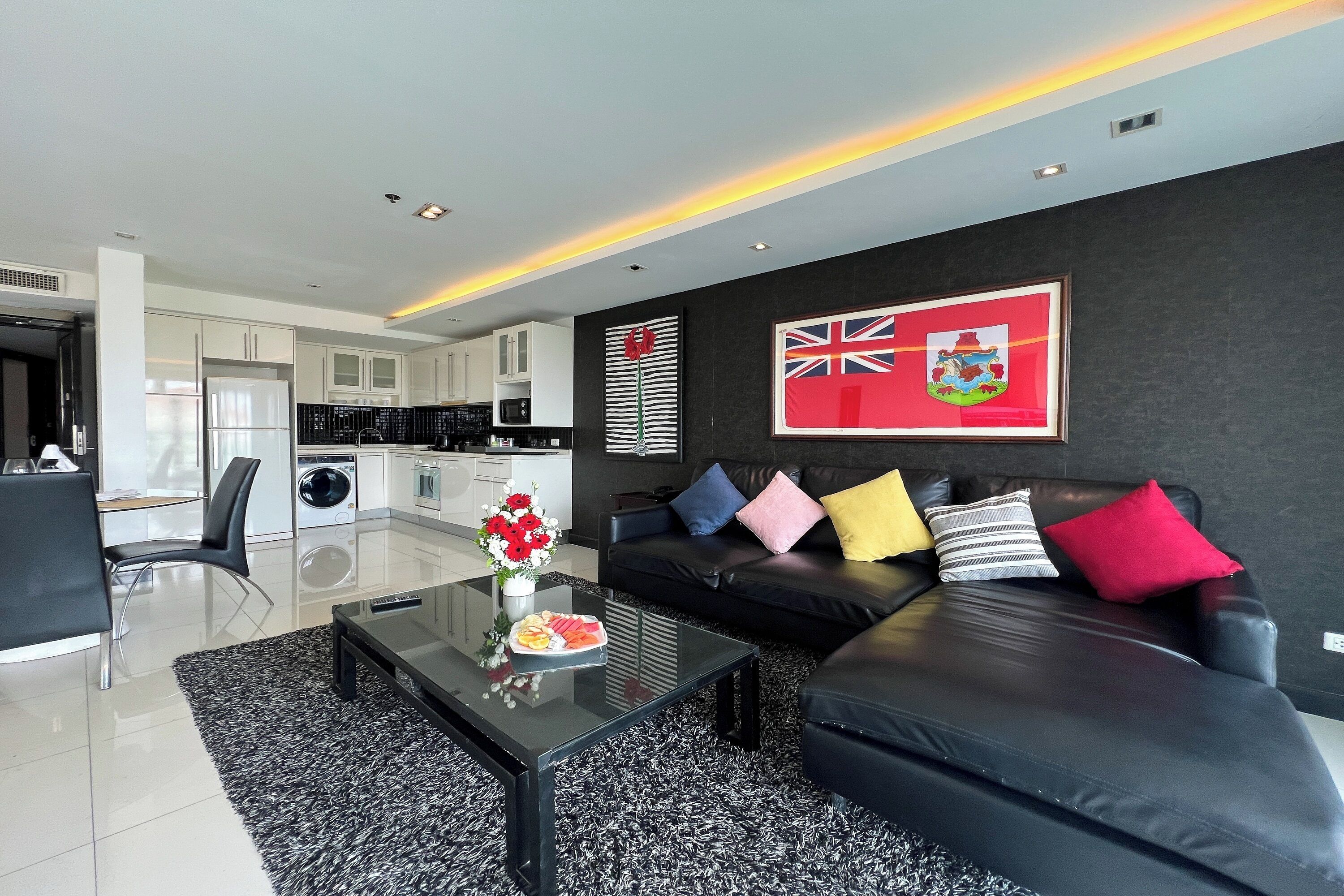A modern living room with a black leather sofa, a glass coffee table, and a dining area with white cabinetry.