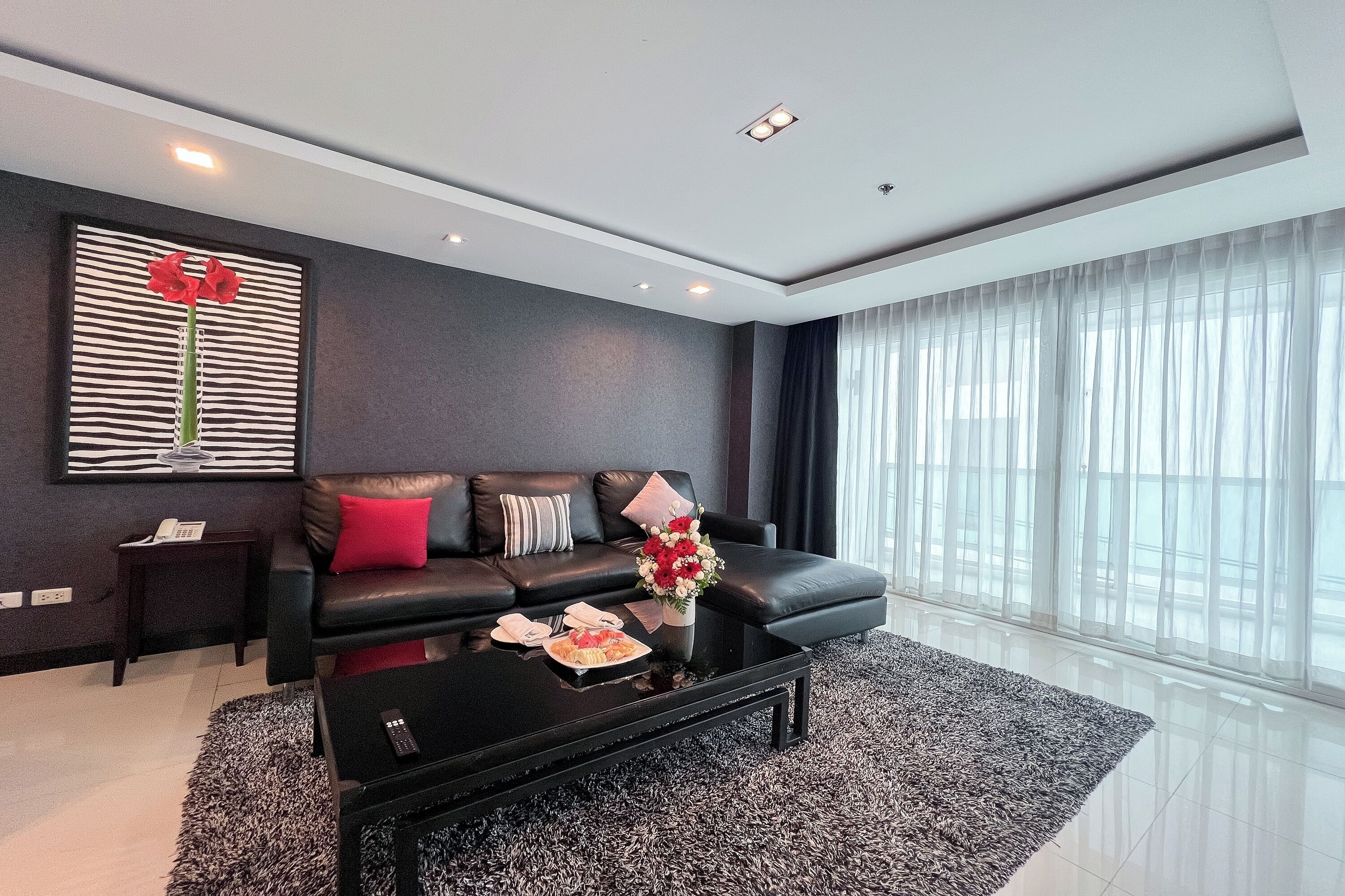 A modern living room with a black leather sofa, a coffee table, and a large window with sheer curtains.