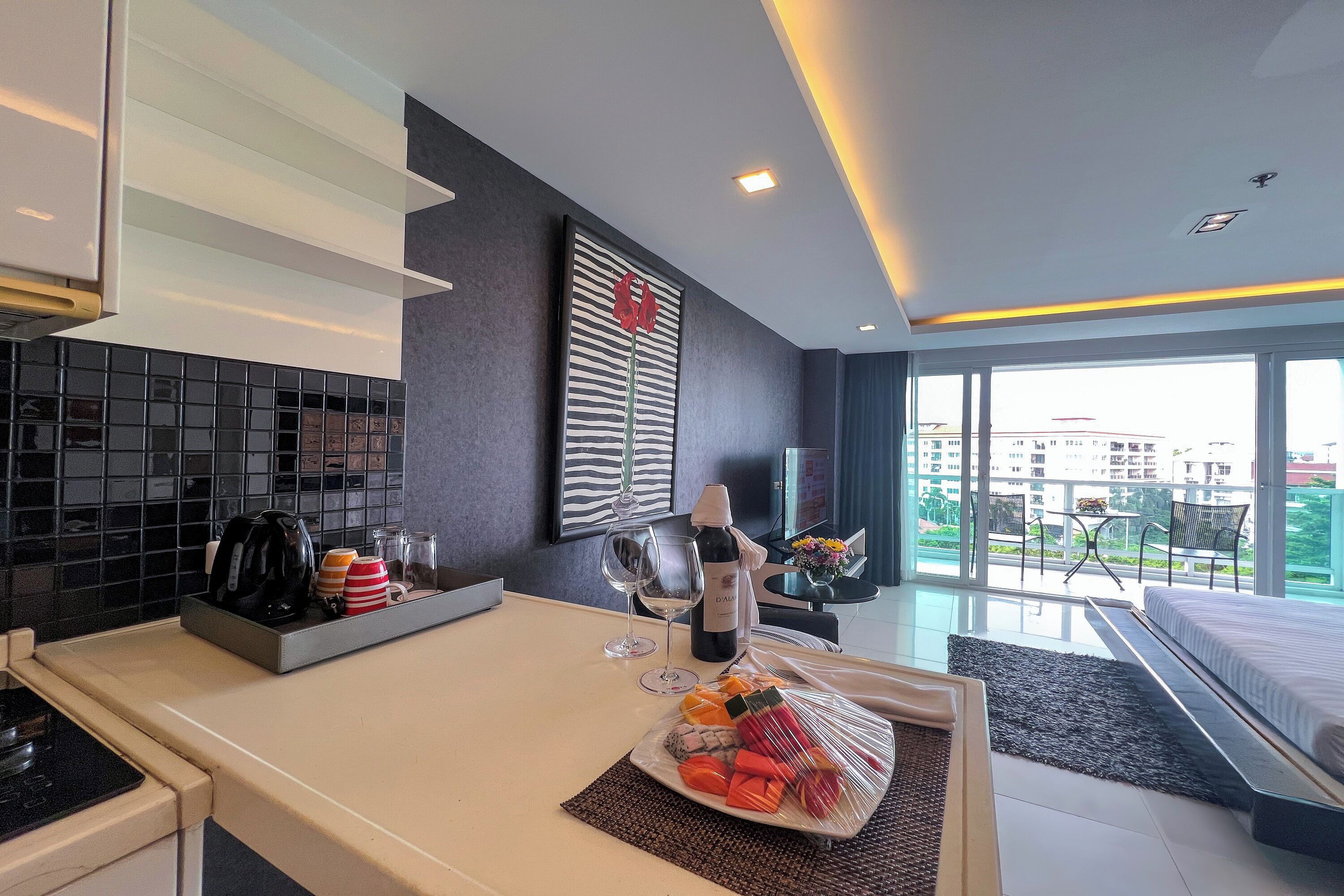 A modern hotel room with a kitchen area, a dining table with a bottle of wine and glasses, and a balcony with a view.