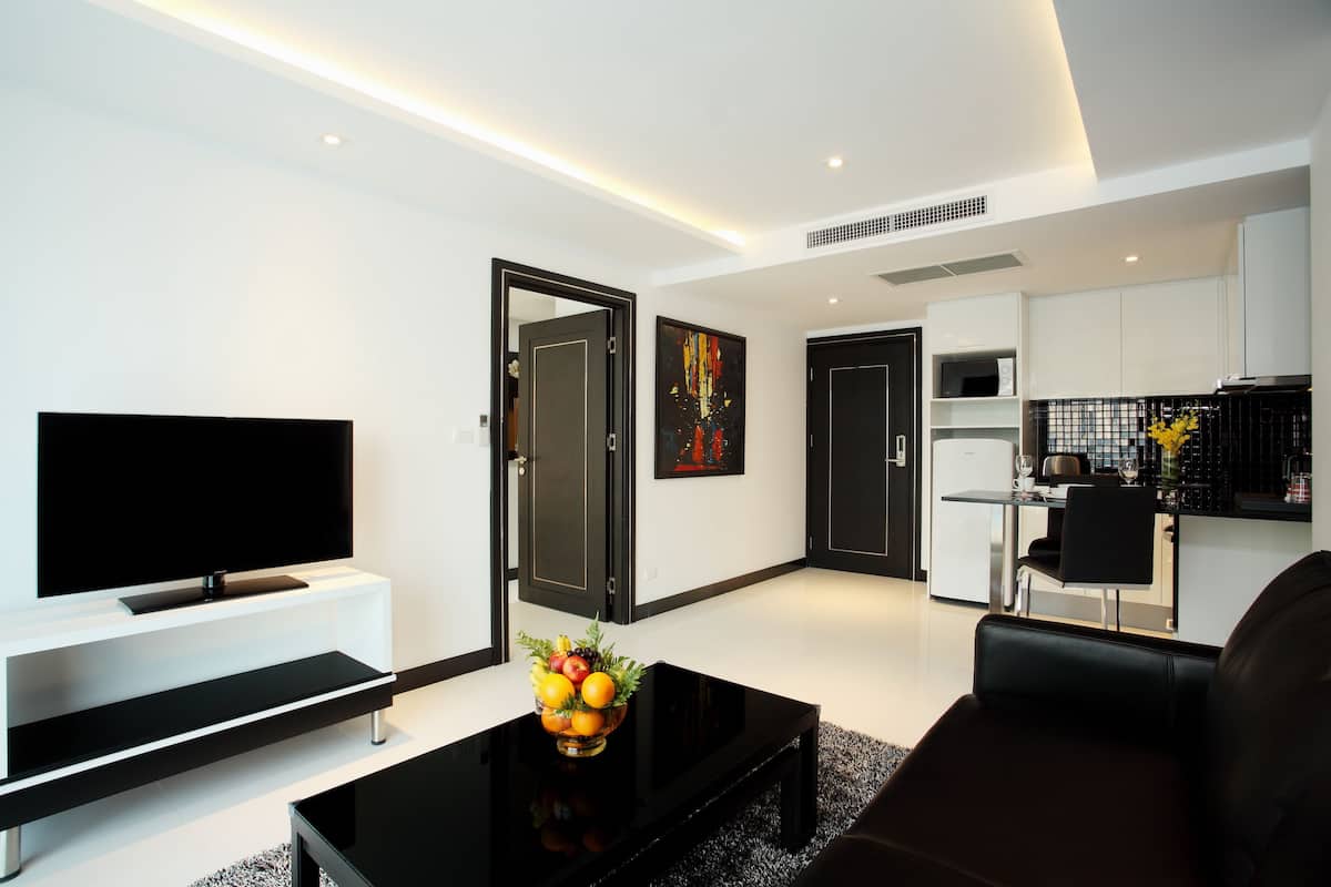Una sala moderna con un televisor de pantalla plana, una mesa de centro negra con fruta, un sofá negro y una zona de cocina con una mesa de comedor y sillas.
