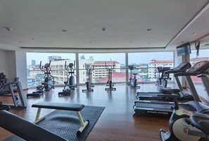 A gym with various exercise equipment, large windows, and a view of buildings. - Nova Suites Pattaya by Compass Hospitality (Pattaya)