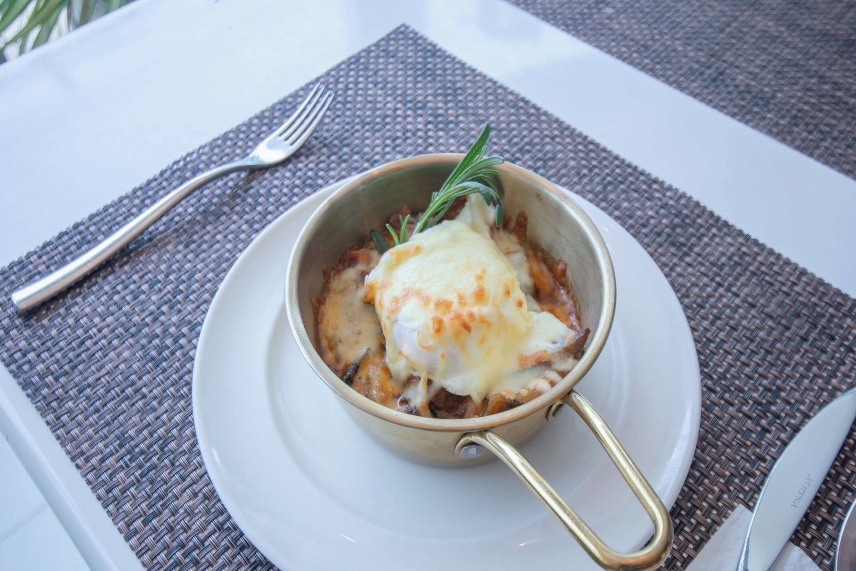 A bowl of food with a handle, placed on a white plate with a fork beside it.