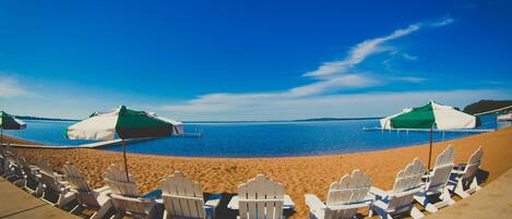 A beach with white Adirondack chairs and green umbrellas, a clear blue sky, and calm water.