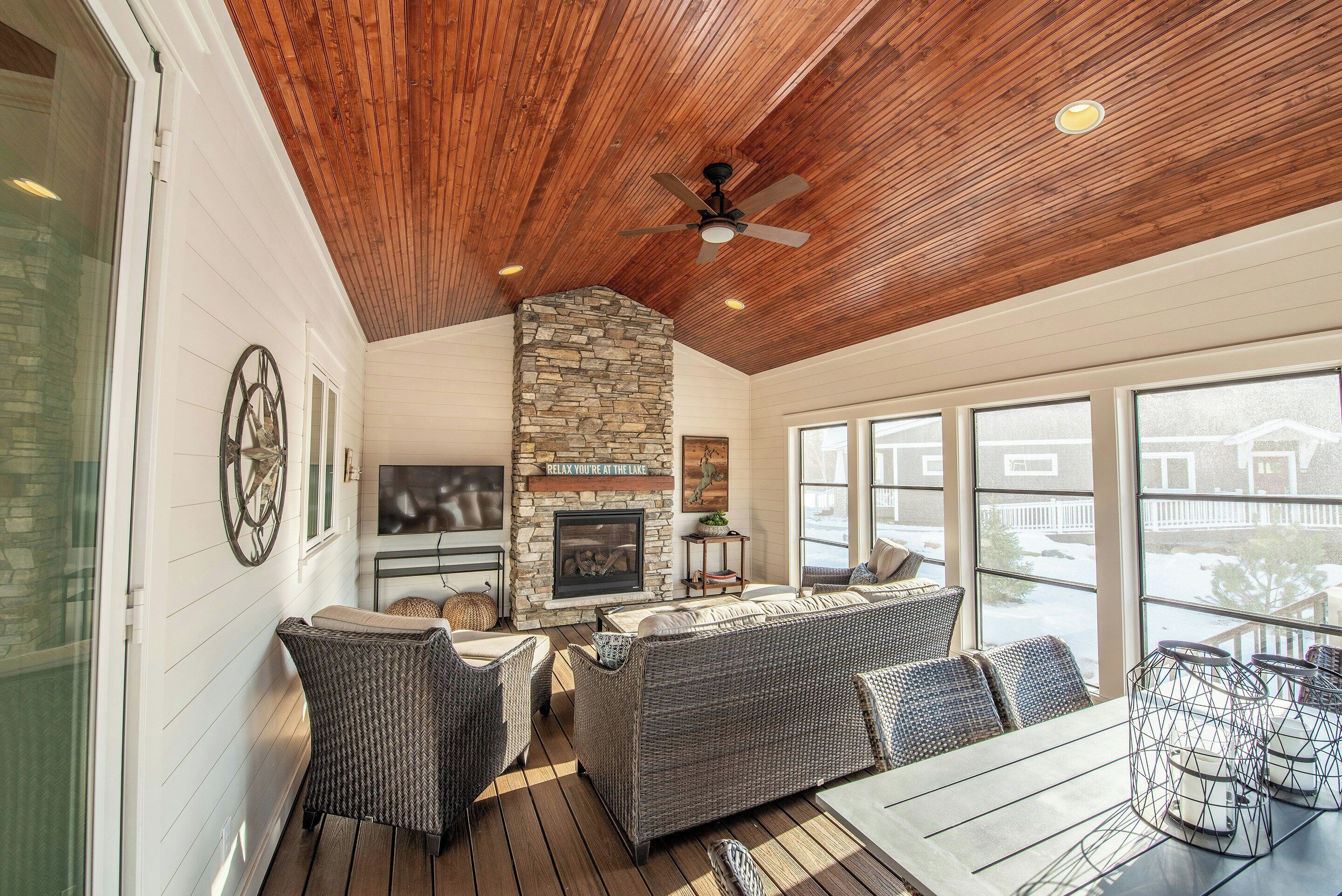 A covered outdoor seating area with wicker furniture, a stone fireplace, and a ceiling fan.