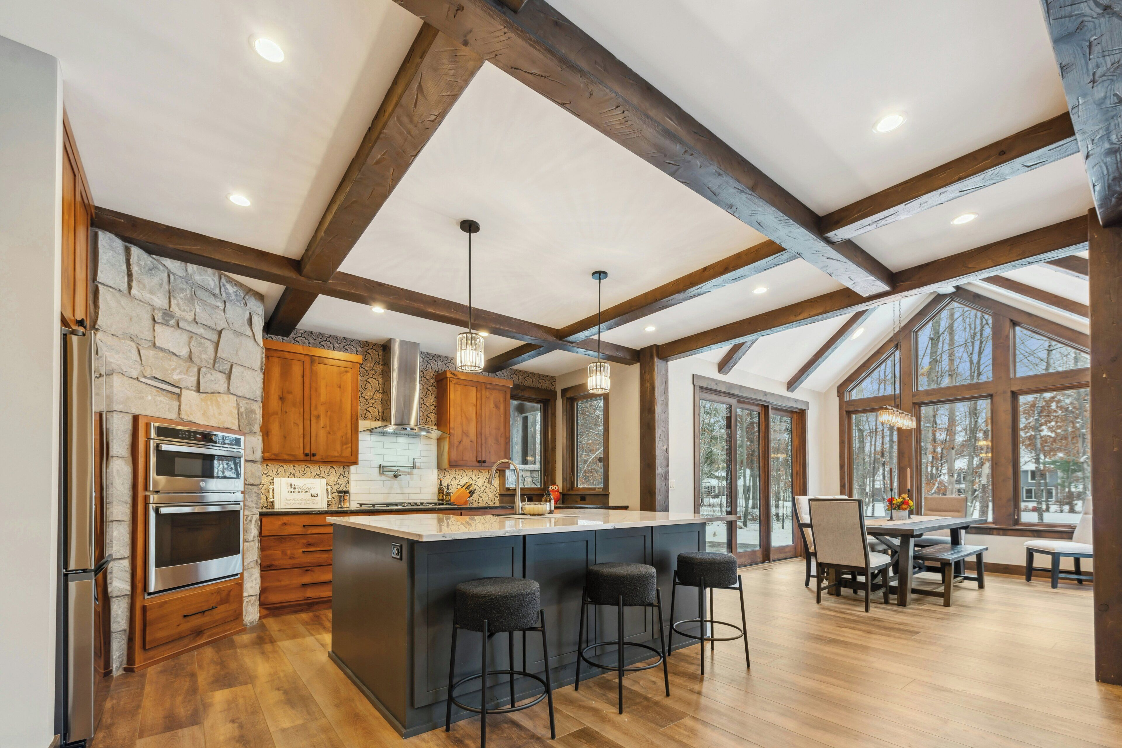 A spacious kitchen with a large island, wooden beams, stone accents, and a view through large windows.