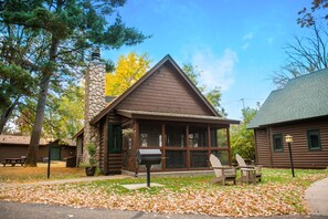 A log cabin with a stone chimney, a porch with chairs, and a mailbox.